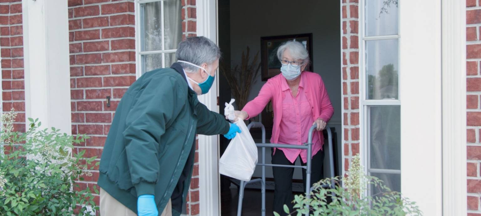 man in gloves and mask handing older woman with walker a bag of food at her front door