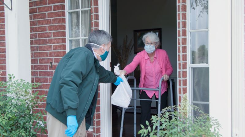 man with gloves and mask handing senior woman with a walker a bag of food at her front door